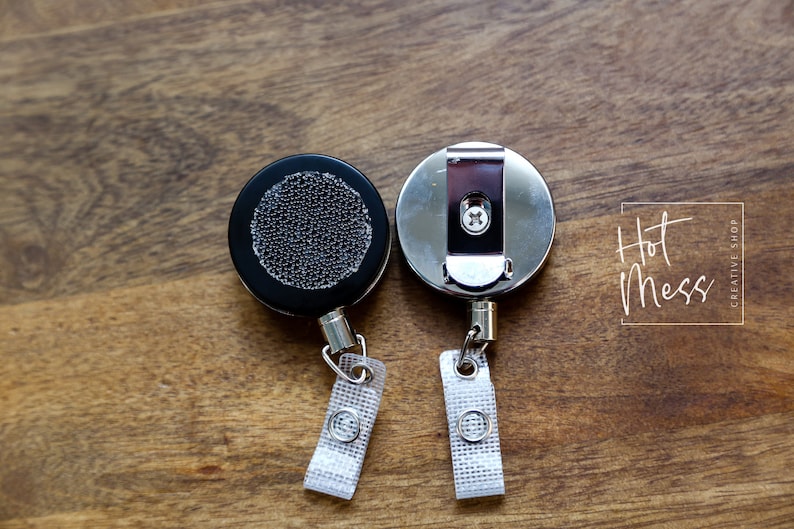 May include: Two retractable badge reels, one with a black plastic case and a textured surface, and the other with a silver metal case. Both reels have a silver metal clip and a white fabric strap.