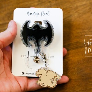Puede incluir: Un carrete de insignia negro y plateado con purpurina con un dise&ntilde;o de murci&eacute;lago negro. El carrete est&aacute; unido a un amuleto de purpurina dorada con una cadena plateada. La tarjeta dice "Badge Reel" y "Hot Mess Creative Shop".