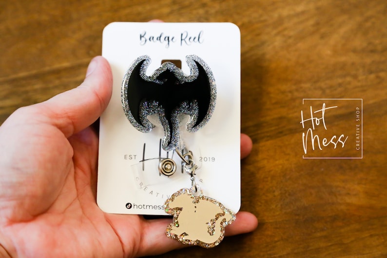 Puede incluir: Un carrete de insignia negro y plateado con purpurina con un dise&ntilde;o de murci&eacute;lago negro. El carrete est&aacute; unido a un amuleto de purpurina dorada con una cadena plateada. La tarjeta dice "Badge Reel" y "Hot Mess Creative Shop".