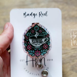 May include: A pink and blue glitter badge reel with a floral design and a teacup with the text "Suriel Tea Co."