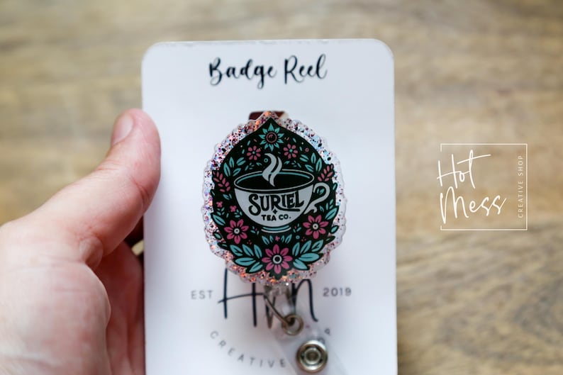 May include: A pink and blue glitter badge reel with a floral design and a teacup with the text "Suriel Tea Co."