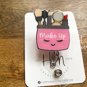 May include: Pink makeup bag with a cute face and eyelashes. The bag has a black outline and the words "Make Up" written on it. The bag is attached to a silver retractable badge reel with a clear plastic strap. The badge reel has the text "Hot Mess Creative Shop" and "Est 2019" on it.
