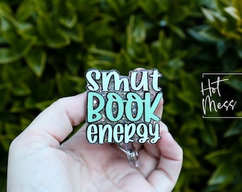 Può includere: Un portabadge in glitter blu turchese e argento con il testo "smut book energy" su di esso. Il portabadge è tenuto da una mano. Il testo "Hot Mess Creative Shop" è visibile sullo sfondo.