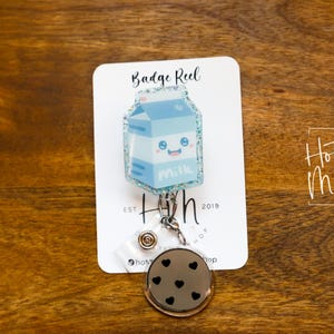 Può includere: Un cartone di latte blu e bianco con un simpatico viso da cartone animato è attaccato a un portabadge retrattile argentato con un ciondolo a pois bianco e nero. Il portabadge è etichettato "Badge Reel" e "Hot Mess" con il testo "Est. 2019".