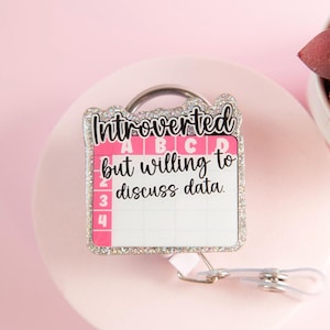 May include: A retractable badge reel with a pink and white design. The badge reel has the words "Introverted but willing to discuss data" in black script. The border is silver glitter. A small pink succulent plant is in the background.