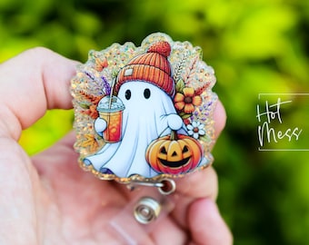 Puede incluir: Un fantasma blanco con un gorro de punto rojo y naranja sostiene una calabaza y una taza de líquido naranja. El fantasma está rodeado de follaje de otoño y purpurina. La imagen es un amuleto o insignia con temática de Halloween.