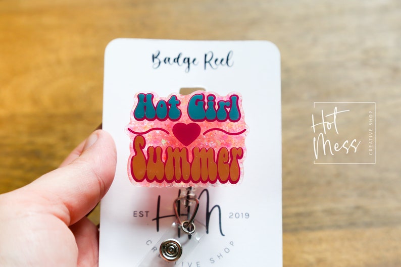 Pode incluir: Um carretel de crach&aacute; de glitter rosa com uma fonte retr&ocirc; turquesa e laranja que diz "Hot Girl Summer" e um cora&ccedil;&atilde;o vermelho. O carretel de crach&aacute; est&aacute; preso a um cart&atilde;o branco com o texto "Badge Reel" e "Hot Mess Creative Shop" impresso nele.