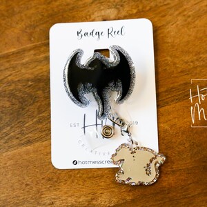 Puede incluir: Un carrete de credencial negro y plateado con purpurina con un dise&ntilde;o de drag&oacute;n. El carrete est&aacute; unido a una cadena plateada con un peque&ntilde;o dije de drag&oacute;n. La tarjeta dice "Badge Reel" y "Hot Mess Creative Shop".