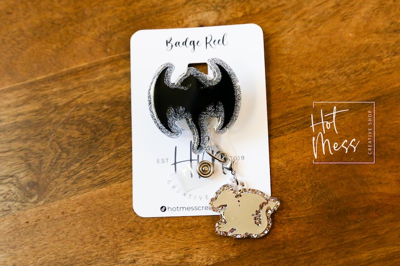 Puede incluir: Un carrete de credencial negro y plateado con purpurina con un dise&ntilde;o de drag&oacute;n. El carrete est&aacute; unido a una cadena plateada con un peque&ntilde;o dije de drag&oacute;n. La tarjeta dice "Badge Reel" y "Hot Mess Creative Shop".