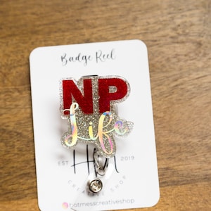 May include: A silver glitter badge reel with a red "NP" and silver glitter "life" text. The reel is attached to a white card with the text "Badge Reel Est 2019 Hot Mess Creative Shop".