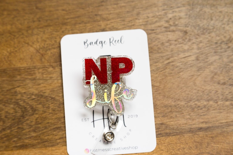 May include: A silver glitter badge reel with a red "NP" and silver glitter "life" text. The reel is attached to a white card with the text "Badge Reel Est 2019 Hot Mess Creative Shop".