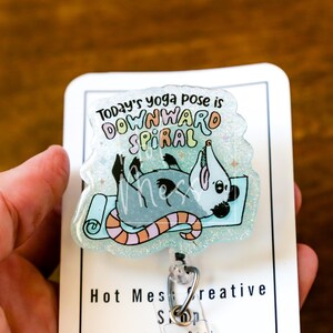 Op de afbeelding: Een badge reel met een cartoon opossum op een yogamat. De tekst luidt "Today's yoga pose is downward spiral". De opossum is grijs met zwarte accenten en de mat is lichtblauw met een gestreept oranje en roze ontwerp.