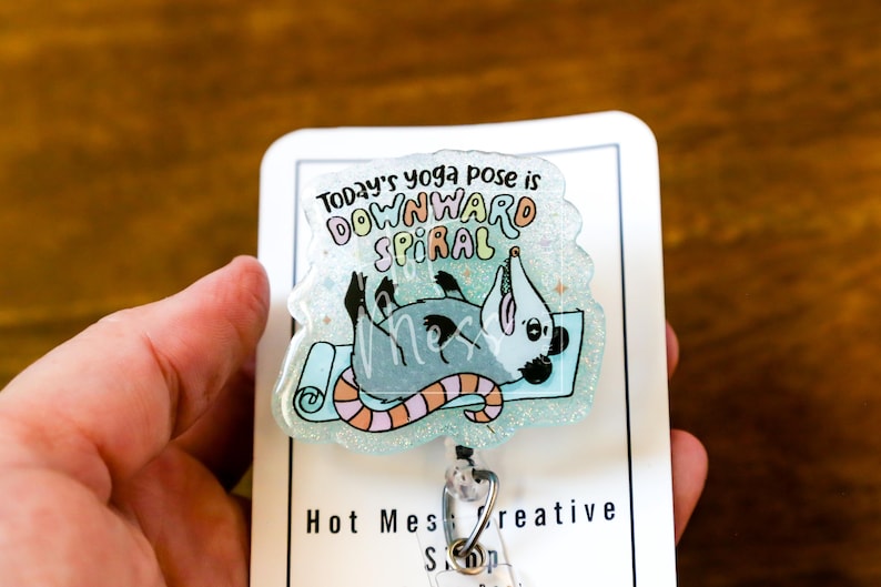 Op de afbeelding: Een badge reel met een cartoon opossum op een yogamat. De tekst luidt "Today's yoga pose is downward spiral". De opossum is grijs met zwarte accenten en de mat is lichtblauw met een gestreept oranje en roze ontwerp.