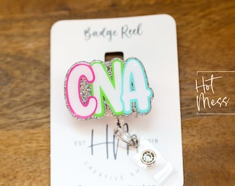May include: A colourful "CNA" badge reel. The letters are outlined in pink, green, and blue, with a silver glitter centre. The badge reel is attached to a white card with the text "Badge Reel" and "Hot Mess Creative Shop".
