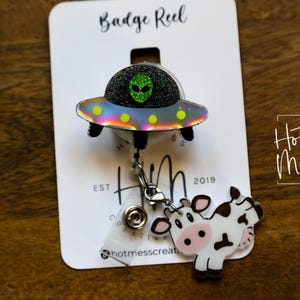 Puede incluir: Un carrete de insignia con un dise&ntilde;o de OVNI y alien&iacute;gena, y un dije de vaca. El OVNI tiene una parte superior de purpurina negra, una cara de alien&iacute;gena verde y una parte inferior iridiscente del arco&iacute;ris. El dije de vaca es blanco con manchas negras y una cara rosa. El carrete de insignia est&aacute; en una tarjeta blanca.