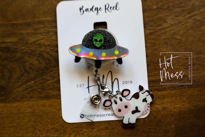 Puede incluir: Un carrete de insignia con un dise&ntilde;o de OVNI y alien&iacute;gena, y un dije de vaca. El OVNI tiene una parte superior de purpurina negra, una cara de alien&iacute;gena verde y una parte inferior iridiscente del arco&iacute;ris. El dije de vaca es blanco con manchas negras y una cara rosa. El carrete de insignia est&aacute; en una tarjeta blanca.