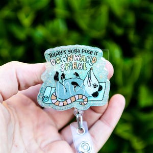 Op de afbeelding: Een heldere, glinsterende badge met een cartoonrat in een yogahouding op een blauwe mat. De tekst "Today's yoga pose is DOWNWARD SPIRAL" staat bovenaan. De badge heeft een doorzichtige plastic clip.