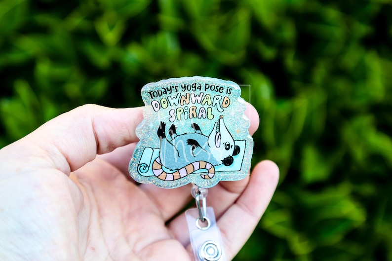 Op de afbeelding: Een heldere, glinsterende badge met een cartoonrat in een yogahouding op een blauwe mat. De tekst "Today's yoga pose is DOWNWARD SPIRAL" staat bovenaan. De badge heeft een doorzichtige plastic clip.