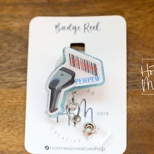 May include: A blue and white badge reel with a cartoon scanner and the text "PEWPEW" on it. The badge reel is on a white card with the text "Badge Reel" and "Hot Mess Creative Shop" on it.