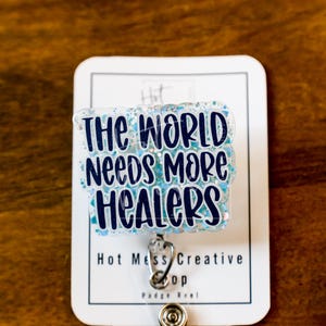 May include: A badge reel with the text "THE WORLD NEEDS MORE HEALERS" in dark blue lettering on a light blue glitter background. The badge reel is attached to a white card with the text "Hot Mess Creative" and "Badge Reel".