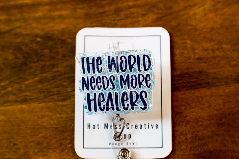 May include: A badge reel with the text "THE WORLD NEEDS MORE HEALERS" in dark blue lettering on a light blue glitter background. The badge reel is attached to a white card with the text "Hot Mess Creative" and "Badge Reel".