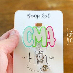 K&ouml;nnte beinhalten: Ein Ausweis-Rollhalter mit den Buchstaben "CMA" in einem farbenfrohen Bubble-Letter-Stil. Die Rolle hat einen Stethoskop-Anh&auml;nger und ist an einer wei&szlig;en Karte mit dem Text "Badge Reel" und "Hot Mess Creative Shop" befestigt.
