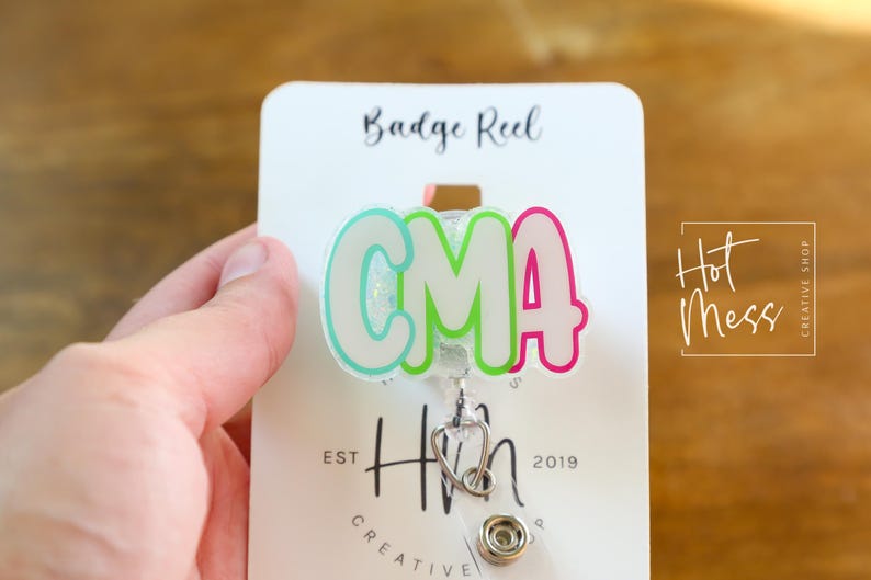 K&ouml;nnte beinhalten: Ein Ausweis-Rollhalter mit den Buchstaben "CMA" in einem farbenfrohen Bubble-Letter-Stil. Die Rolle hat einen Stethoskop-Anh&auml;nger und ist an einer wei&szlig;en Karte mit dem Text "Badge Reel" und "Hot Mess Creative Shop" befestigt.