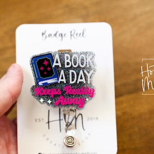 May include: A silver glitter badge reel with a pink and blue book charm. The charm says "A book a day keeps reality away".