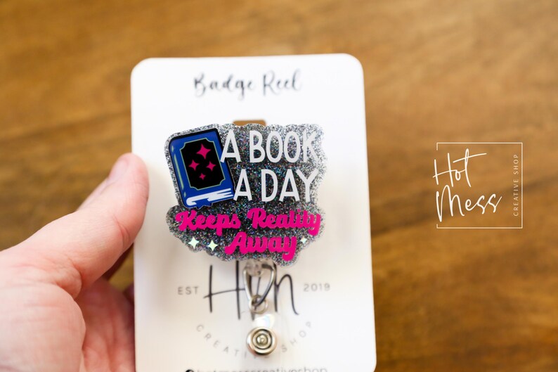 May include: A silver glitter badge reel with a pink and blue book charm. The charm says "A book a day keeps reality away".