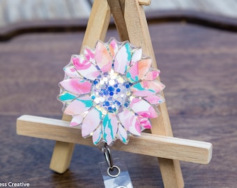 Puede incluir: Un carrete de identificación con estampado floral rosa y verde con purpurina y detalles azules. El carrete está unido a un porta-identificaciones de plástico transparente.