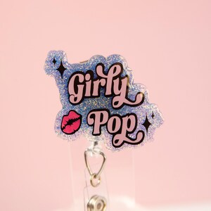 Op de afbeelding: Een glitterende badge reel met de woorden "Girly Pop" in roze en zwarte letters. De badge reel heeft een lichtblauwe glitterachtergrond met een roze lipafbeelding en zwarte steraccenten. De reel heeft een transparant intrekbaar plastic koord.
