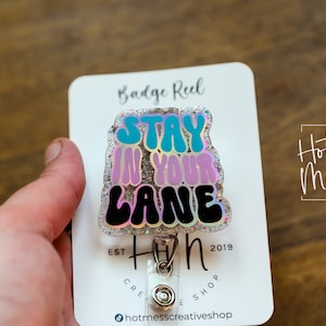 May include: A silver glitter badge reel with a holographic finish. The reel features a black, pink, and blue graphic that says "Stay in your lane".
