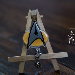 May include: A yellow and gray Star Trek insignia badge reel with a clear plastic casing. The badge reel is on a small wooden easel. The text "Hot Mess Creative Shop" is visible in the background.