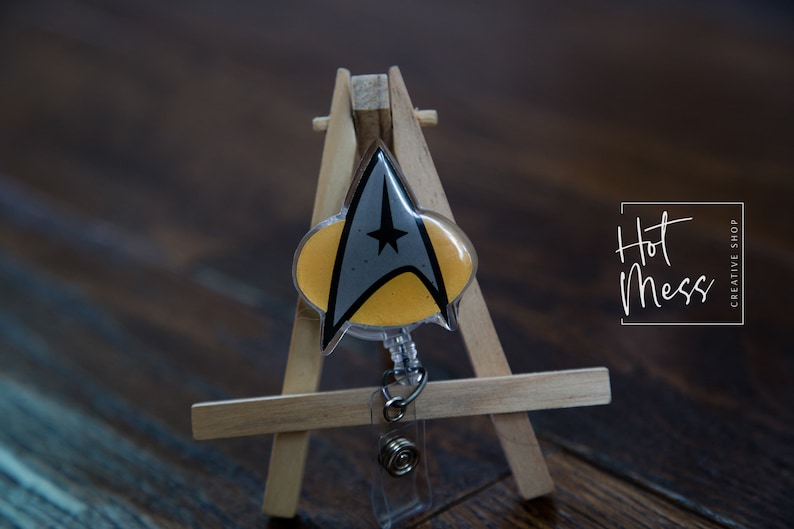 May include: A yellow and gray Star Trek insignia badge reel with a clear plastic casing. The badge reel is on a small wooden easel. The text "Hot Mess Creative Shop" is visible in the background.