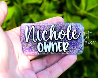 以下が含まれることがあります： 紫色の虹色のグリッターでできたネームタグで、白い文字で「Nichole Owner」と書かれています。