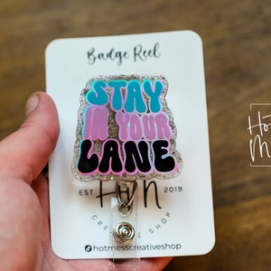 May include: A silver badge reel with a glitter background and a colourful, glittery,  "Stay in your lane"  sticker. The badge reel is attached to a clear plastic retractable cord.