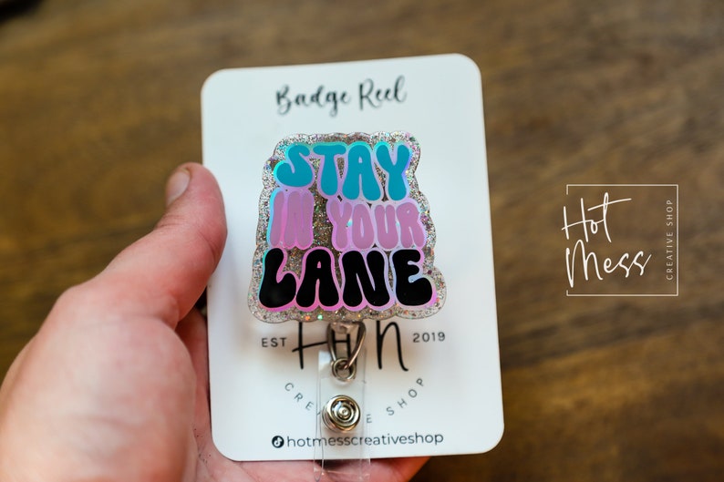 May include: A silver badge reel with a glitter background and a colourful, glittery,  "Stay in your lane"  sticker. The badge reel is attached to a clear plastic retractable cord.