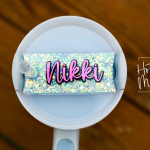 Puede incluir: Un vaso blanco con una tapa azul y una pegatina de brillantina que dice "Nikki". La pegatina está en la tapa del vaso. El vaso está sobre una superficie de madera.