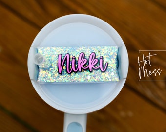 Puede incluir: Un vaso blanco con una tapa azul y una pegatina de brillantina que dice "Nikki". La pegatina está en la tapa del vaso. El vaso está sobre una superficie de madera.