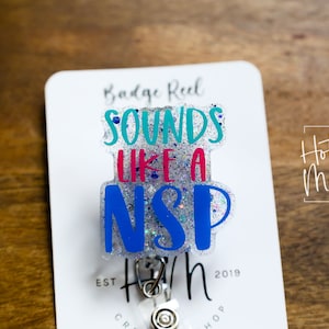Könnte beinhalten: Ein weißer Badge-Rollenhalter mit einem durchsichtigen, glitzernden, blauen, rosa und türkisfarbenen Badge-Halter, auf dem "Sounds Like a  NSP" steht. Der Badge-Rollenhalter befindet sich auf einer weißen Karte mit dem aufgedruckten Text "Badge Reel" und "Hot Mess Creative Shop".