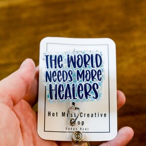 May include: A white badge reel with the text "THE WORLD NEEDS MORE HEALERS" in blue lettering. The badge reel has a clear, glittery overlay and a silver retractable cord clip. The badge reel is held in a person's hand.