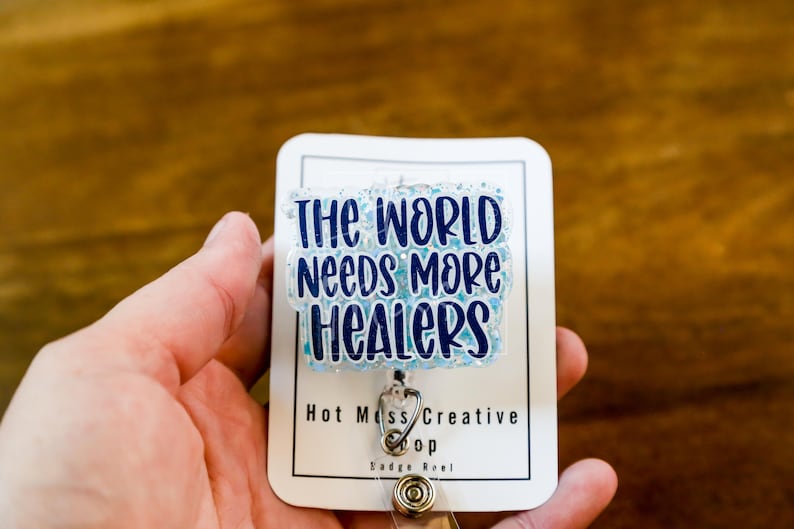 May include: A white badge reel with the text "THE WORLD NEEDS MORE HEALERS" in blue lettering. The badge reel has a clear, glittery overlay and a silver retractable cord clip. The badge reel is held in a person's hand.