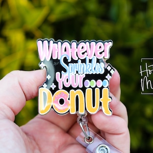 May include: A black and yellow badge holder with the text "Whatever sprinkles your donut" in pink, blue, and yellow. The donut is pink with white frosting and colorful sprinkles. The badge holder has a clear plastic retractable reel.