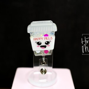 May include: A clear acrylic badge reel featuring a cartoon pill bottle design. The bottle has a white base with the words "HAPPY PILLS" in pink, and a smiling face. The top is a clear lid filled with glitter.