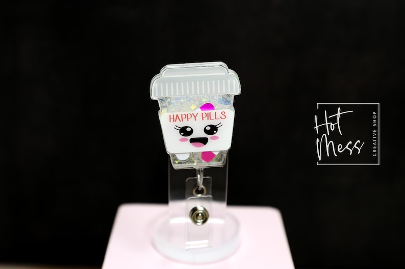 May include: A clear acrylic badge reel featuring a cartoon pill bottle design. The bottle has a white base with the words "HAPPY PILLS" in pink, and a smiling face. The top is a clear lid filled with glitter.