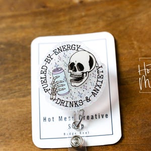 May include: A badge reel with a circular design featuring a skull holding a can, surrounded by text that reads "Fueled by Energy Drinks & Anxiety." The badge reel is on a white card with the shop's name and logo.