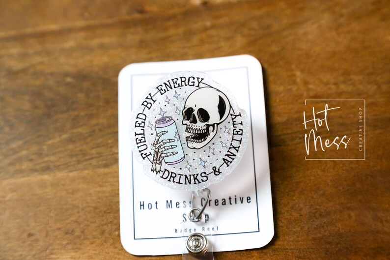 May include: A badge reel with a circular design featuring a skull holding a can, surrounded by text that reads "Fueled by Energy Drinks & Anxiety." The badge reel is on a white card with the shop's name and logo.