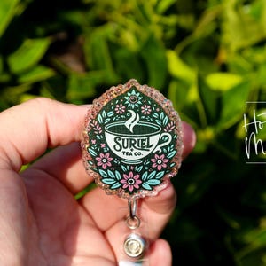 May include: A pink and green glitter badge reel with a teacup design. The teacup has a white background with the text "Suriel Tea Co." in black. The teacup is surrounded by pink and green flowers.