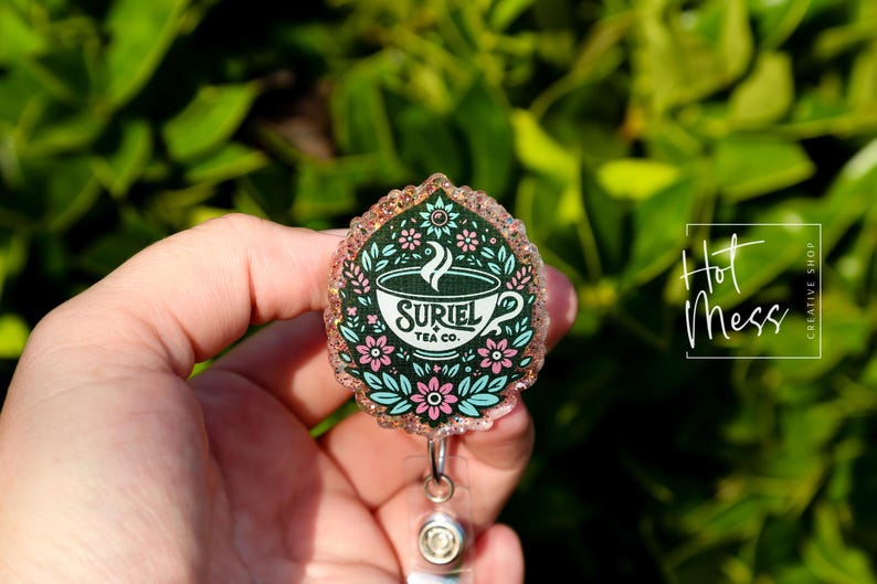 May include: A pink and green glitter badge reel with a teacup design. The teacup has a white background with the text "Suriel Tea Co." in black. The teacup is surrounded by pink and green flowers.