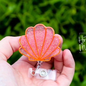 Peut inclure: Un porte-badge rétractable en plastique transparent avec un charme en forme de coquillage en paillettes rose et orange. Le charme est en forme de coquillage avec une surface texturée. Le porte-badge a un clip argenté et une sangle en plastique transparent.
